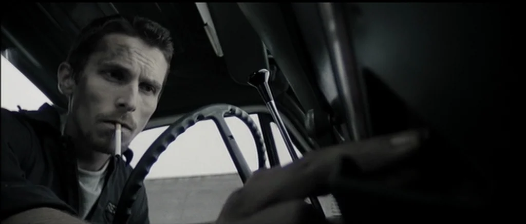 The Machinist (2004) movie still: medium — The interior of a vehicle shows a man sitting in the driver's seat. He smokes a cigarette while resting…