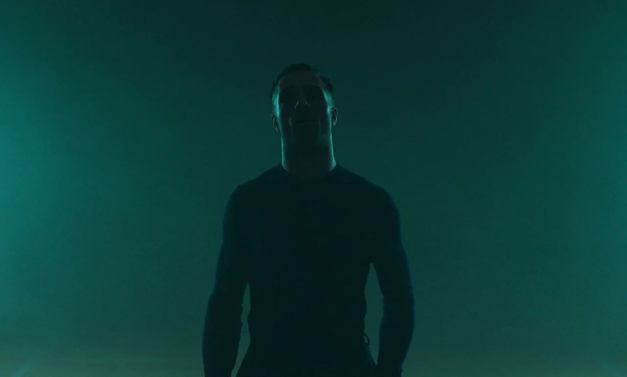 It Is In Us All (2022) movie still: medium — A dimly lit, hazy interior space with a solid dark teal background. A man stands frontally in the…