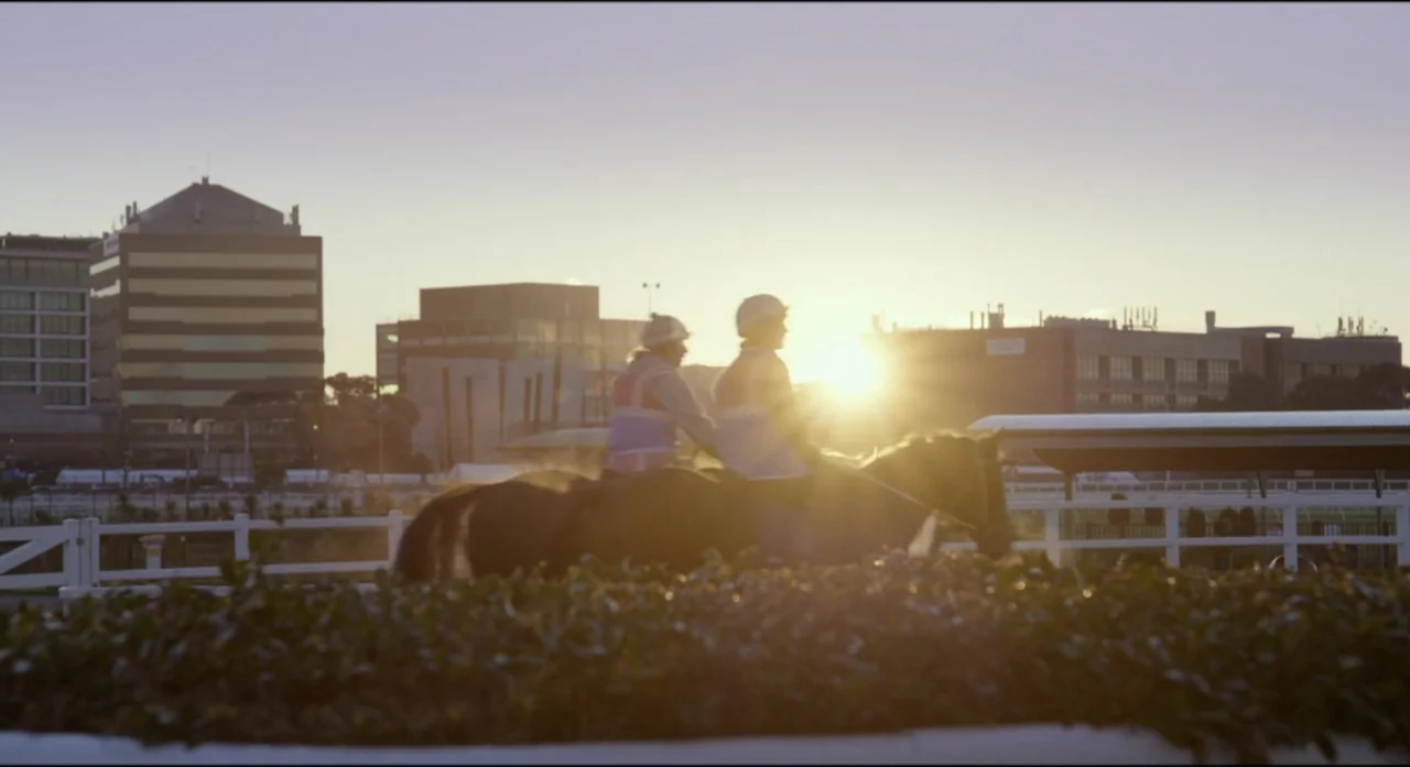Ride Like a Girl (2019) movie still: medium — A horse racing track at golden hour features buildings in the far background. Two riders on horses…