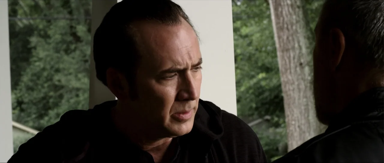 Rage (2014) movie still: closeup — An outdoor porch with a white pillar and dense green foliage in the background. A middle-aged man in a black…