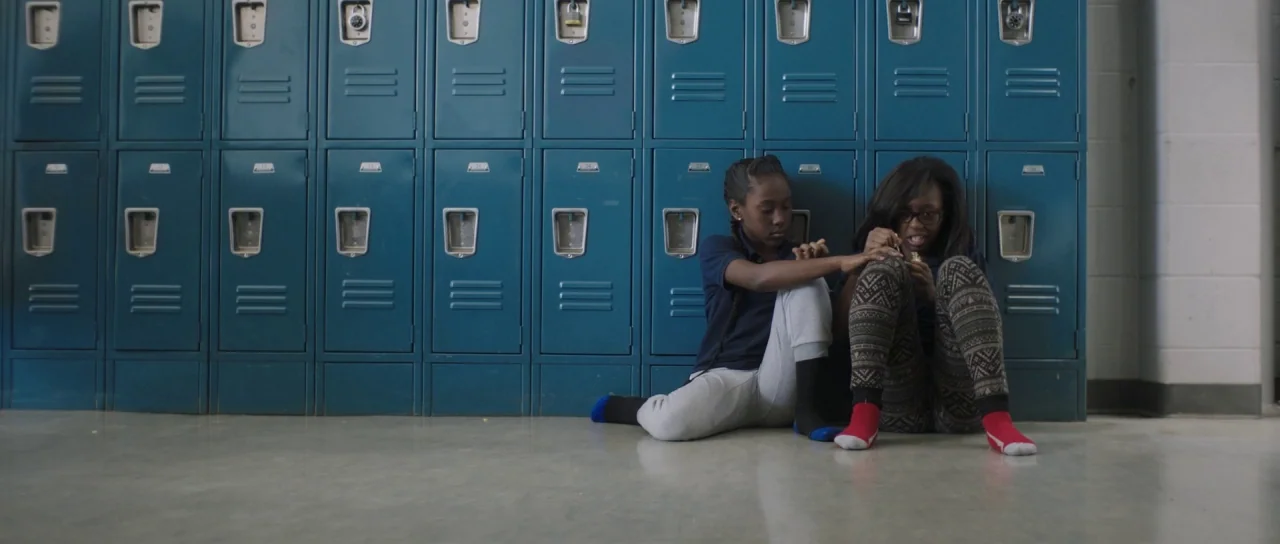 The Fits (2016) movie still: medium — A row of teal metal lockers lines the wall of a school hallway. Two girls sit on the concrete floor while…