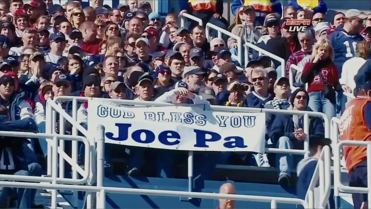 Paterno (2018) movie still: wide — A dense crowd of people sits in blue stadium bleachers under daylight. A white banner reading God Bless You Joe…