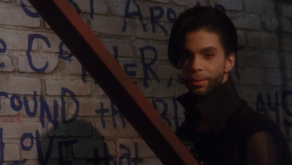 Graffiti Bridge (1990) movie still: medium — A brick wall with blue spray-painted text serves as a backdrop in an alley. Prince stands in front of…