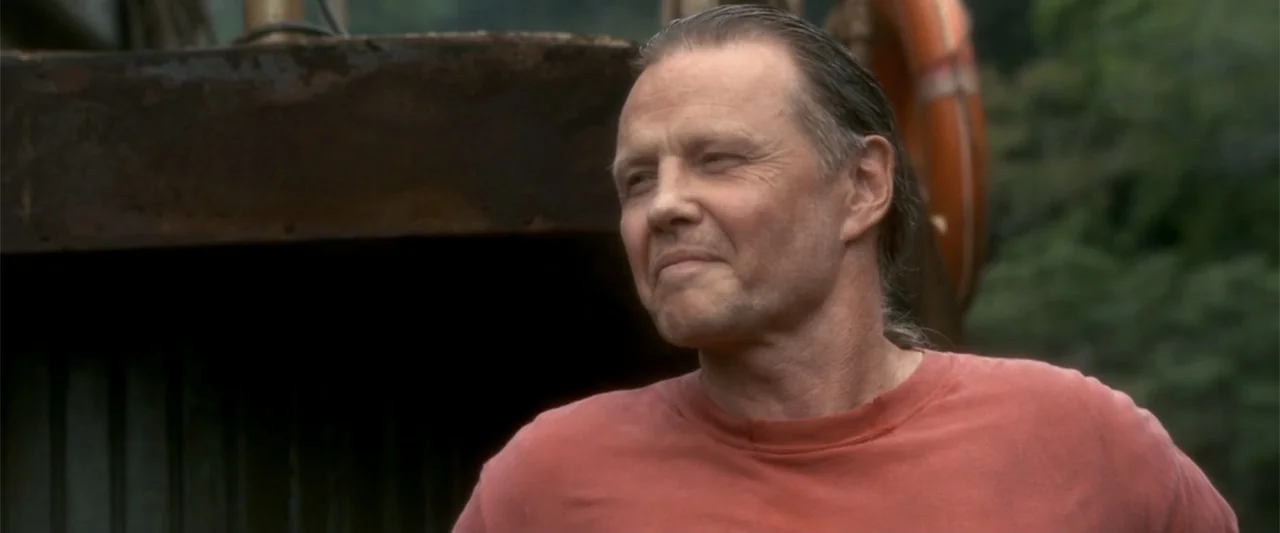 Anaconda (1997) movie still: medium — A man stands on a weathered boat deck near a life ring. He looks toward the side while wearing a simple…