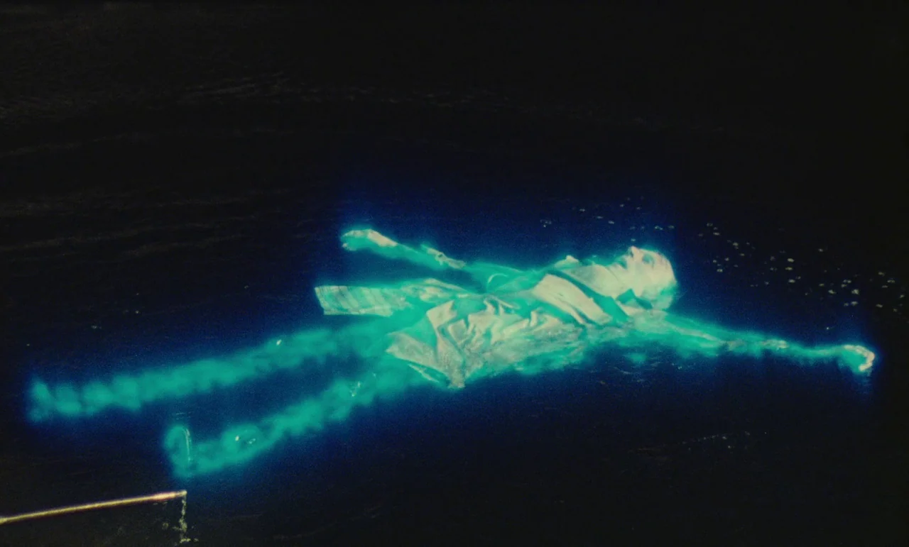 Litan (1982) movie still: wide — A person floats supine on the surface of a dark body of water. The subject is surrounded by a glowing cyan outline…