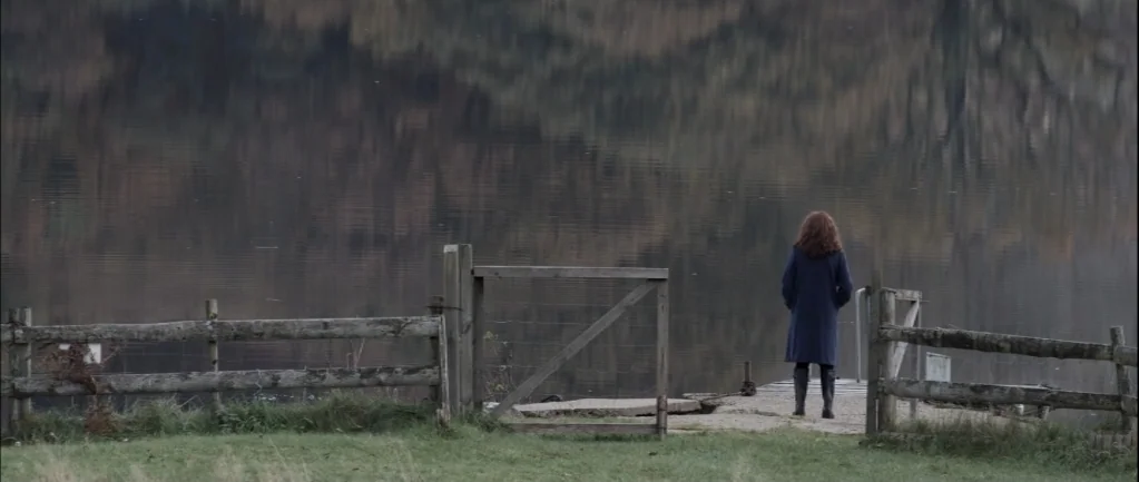 One Hundred Mornings (2009) movie still: fullbody — A dark lake stretches out behind a weathered wooden fence and open gate. A woman stands on a…