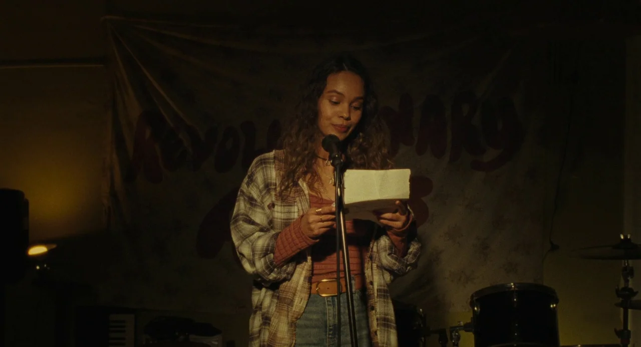 When You Finish Saving The World (2022) movie still: medium — A dimly lit basement room features a wall banner with red text behind the performer. A…