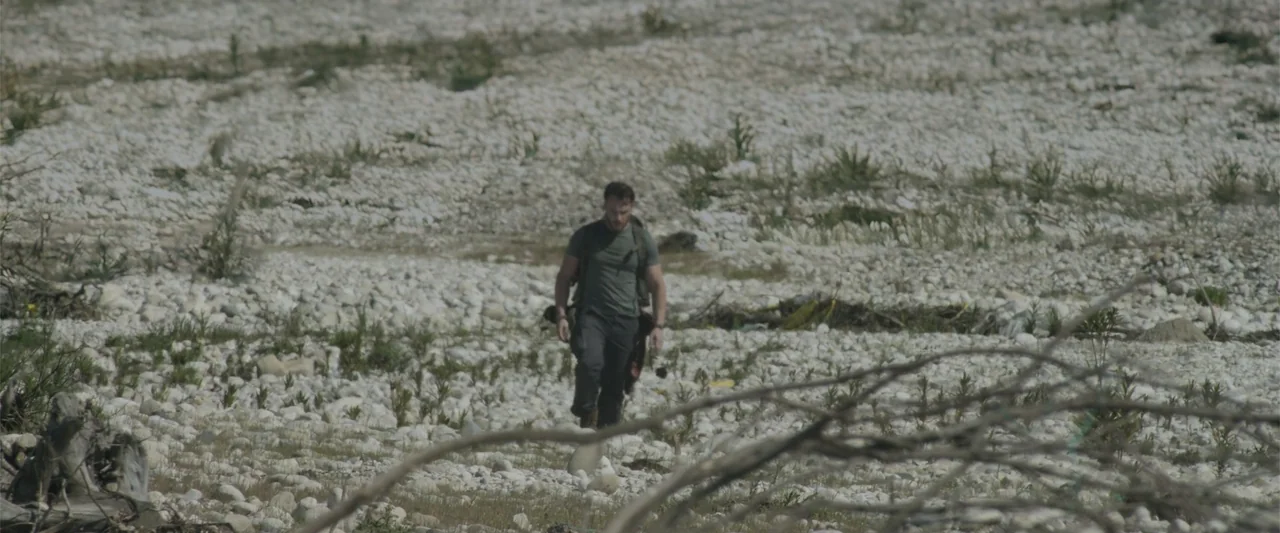Blue Cave (2024) movie still: wide — A wide, dry, white-rock riverbed spans the frame with sparse dry vegetation. A single man walks toward the…