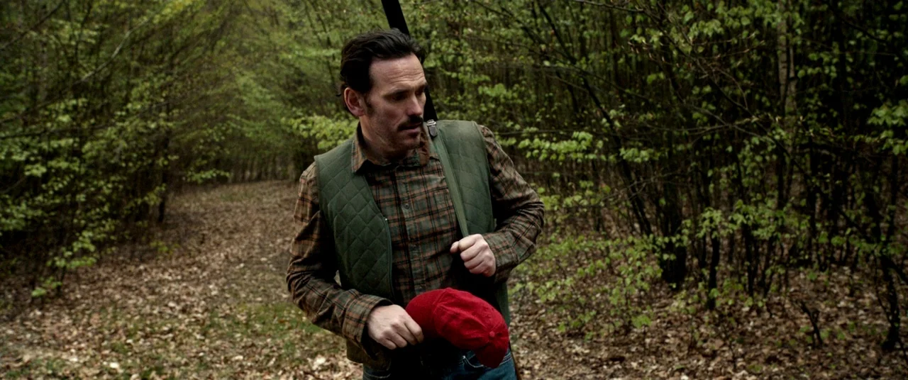 The House That Jack Built (2018) movie still: medium — A man walks along a narrow trail surrounded by dense, leaf-covered deciduous trees. He holds…