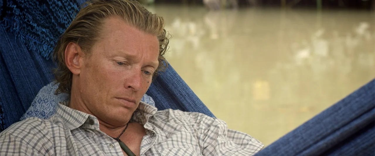 Dirty Little Secret (1998) movie still: medium — A man rests in a blue fabric hammock suspended over murky, still water. He is lying in the hammock…