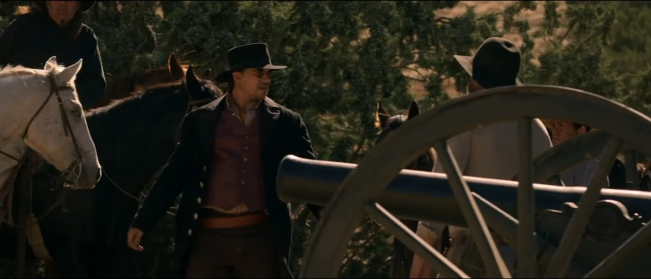 Dead for a Dollar (2022) movie still: medium — A dry, sunlit outdoor area with scrubby trees and a heavy iron field cannon. Two men in western…