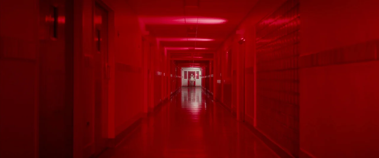 Scary Stories to Tell in the Dark (2019) movie still: wide — A long symmetrical institutional corridor is bathed in intense red light. A lone figure…