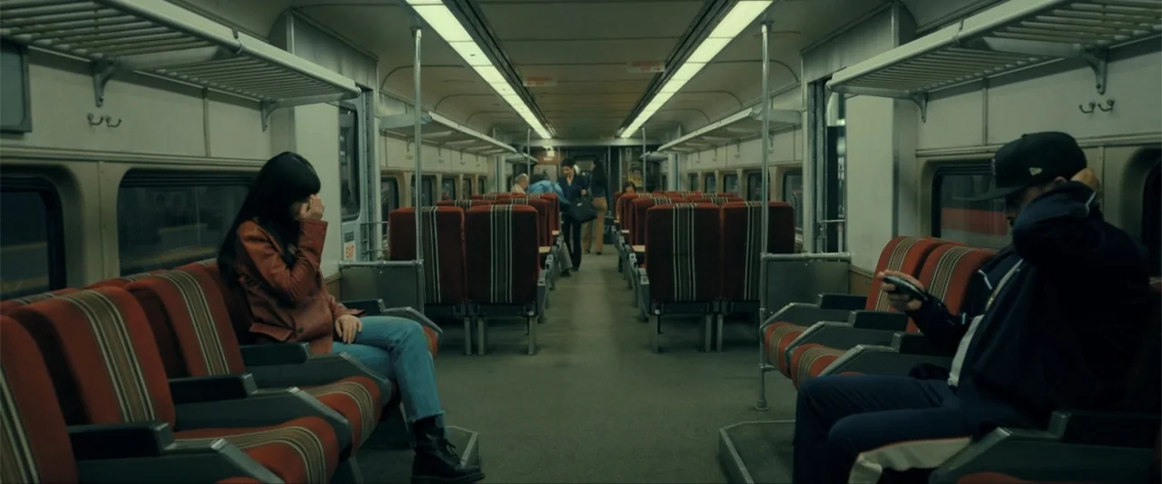 Madame Web (2024) movie still: wide — The interior of a commuter train features rows of striped red upholstered seats. A woman sits on the left…