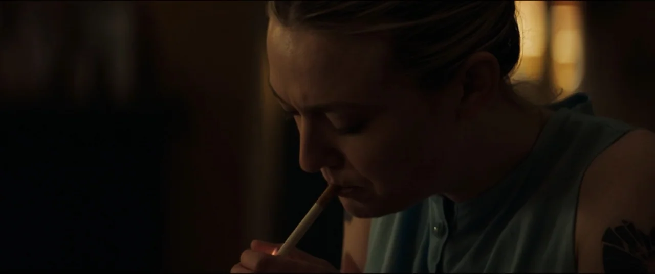 Vicious (2025) movie still: closeup — A dark indoor space with blurred background details visible in the gloom. A woman lights a cigarette with a…