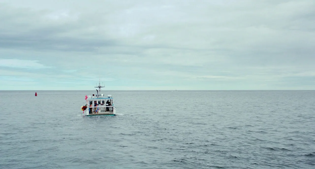 Manchester By The Sea (2016) movie still: wide — A small white fishing boat floats on the vast, calm surface of the sea. The horizon line is visible…