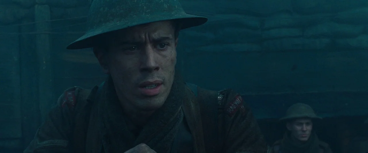 War Horse (2011) movie still: medium — A dark muddy trench with sandbag walls under a dim overcast sky. A young soldier wearing a helmet looks…