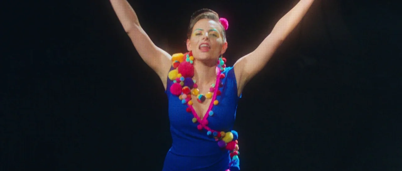 Greener Grass (2019) movie still: medium — A performer stands centered in front of a dark, non-descript background. The woman wears a bright blue…