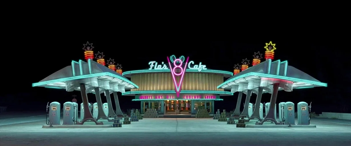Cars (2006) movie still: establishing — A 1950s-style cafe and fuel station is adorned with glowing neon signs at night. The structure features…