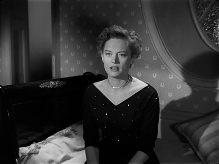 The Sleeping Tiger (1954) movie still: medium — A woman sits on a bed in a room with patterned wallpaper and dark furniture. She looks forward while…
