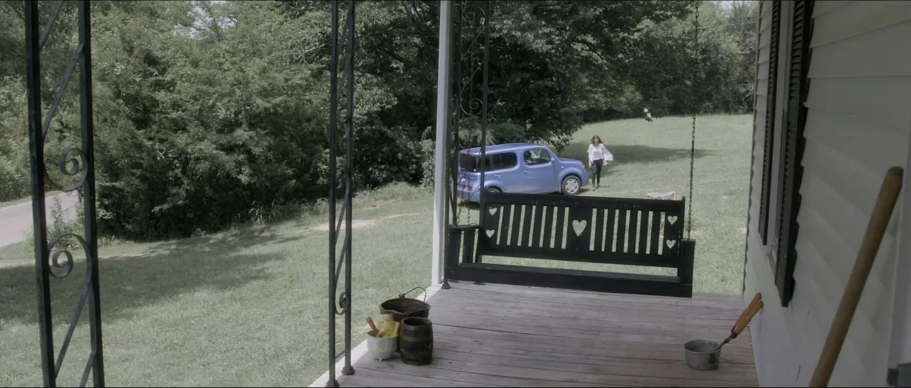 If You're Gone (2019) movie still: wide — A wooden porch with a black swinging bench overlooks a grassy yard. A woman approaches the house across…
