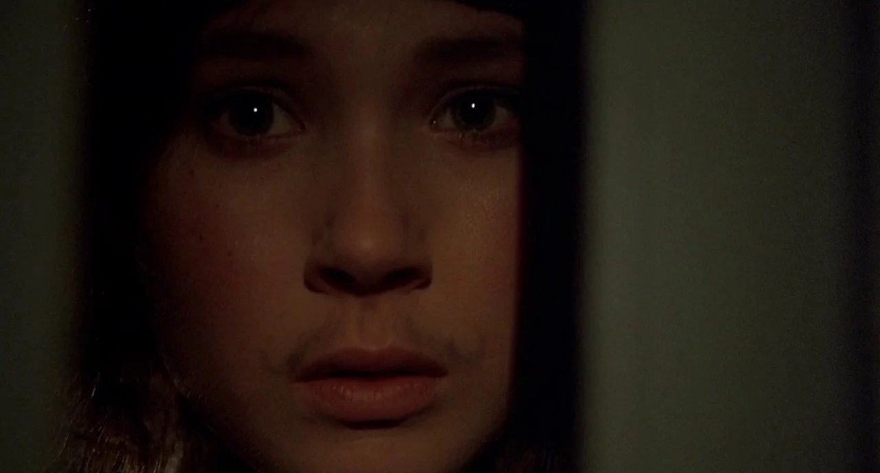 Cracks (2009) movie still: closeup — A dimly lit interior space with the subject's face emerging from deep shadow. The subject gazes directly…