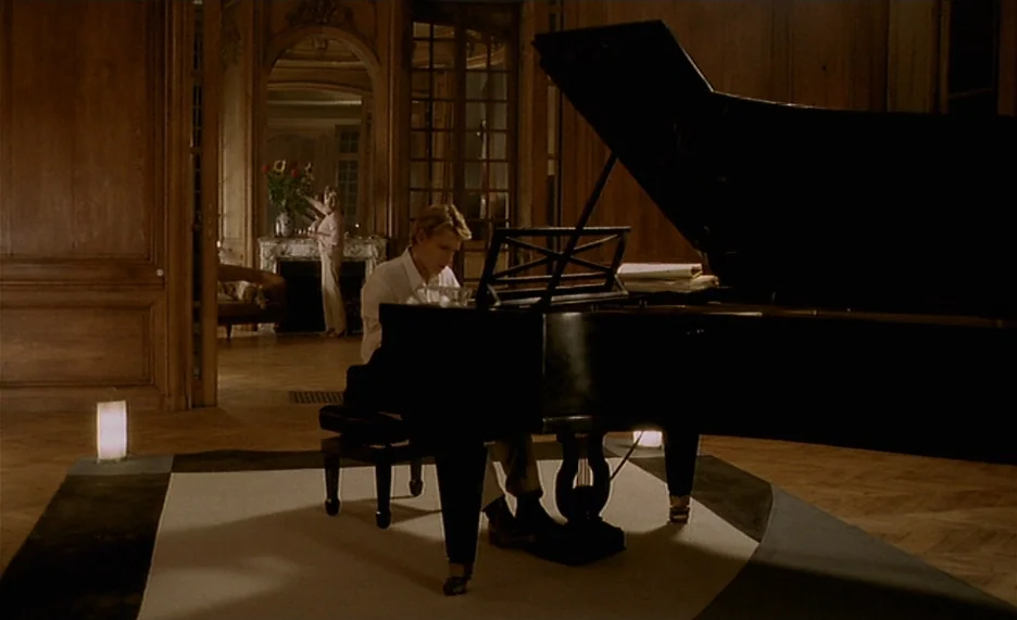 Pola X (1999) movie still: medium — An opulent room with polished wooden wall paneling and a large, dark grand piano. A man plays the piano in the…