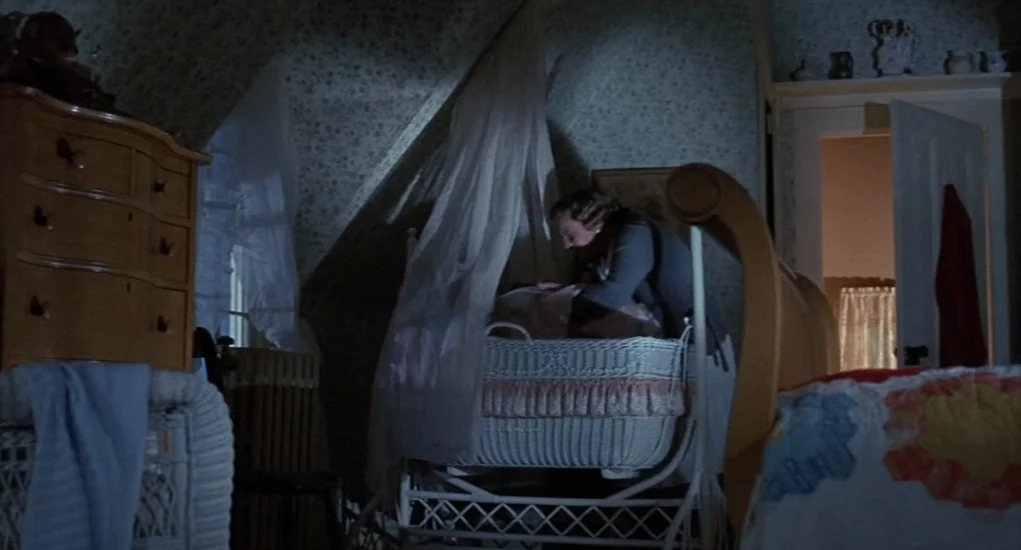 The Other (1972) movie still: medium — A dimly lit nursery features floral wallpaper and a wicker crib under a canopy. A woman in a dark cardigan…