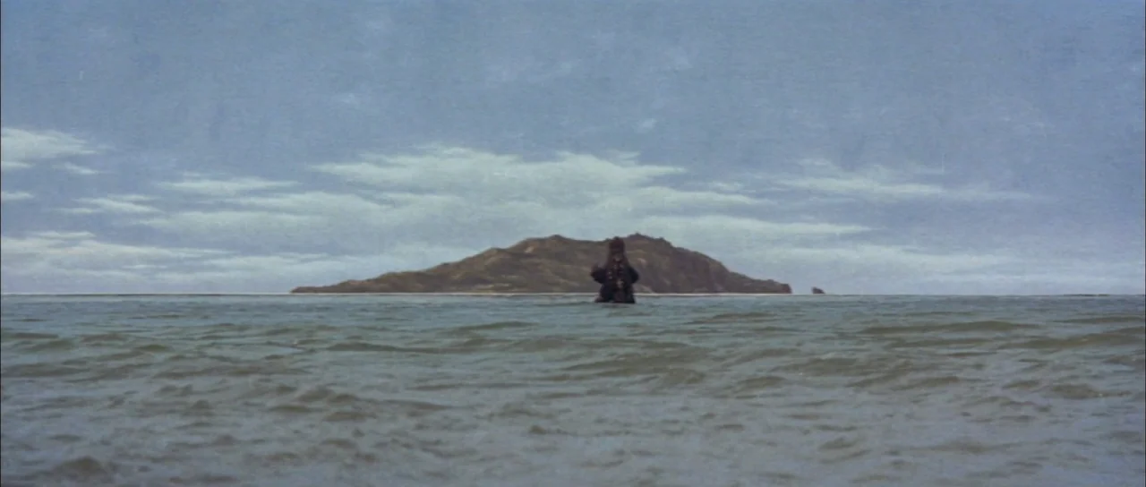 Mothra Vs Godzilla (1964) movie still: wide — A vast, calm ocean stretches toward a low, grassy island under a cloudy sky. The dark figure of a…