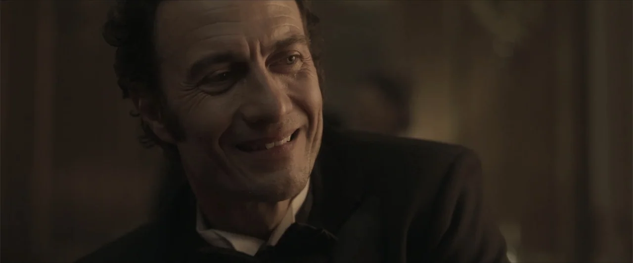 Angela (2023) movie still: closeup — A dimly lit room with muted earth tones and period-appropriate formal attire. A middle-aged man in a tuxedo…
