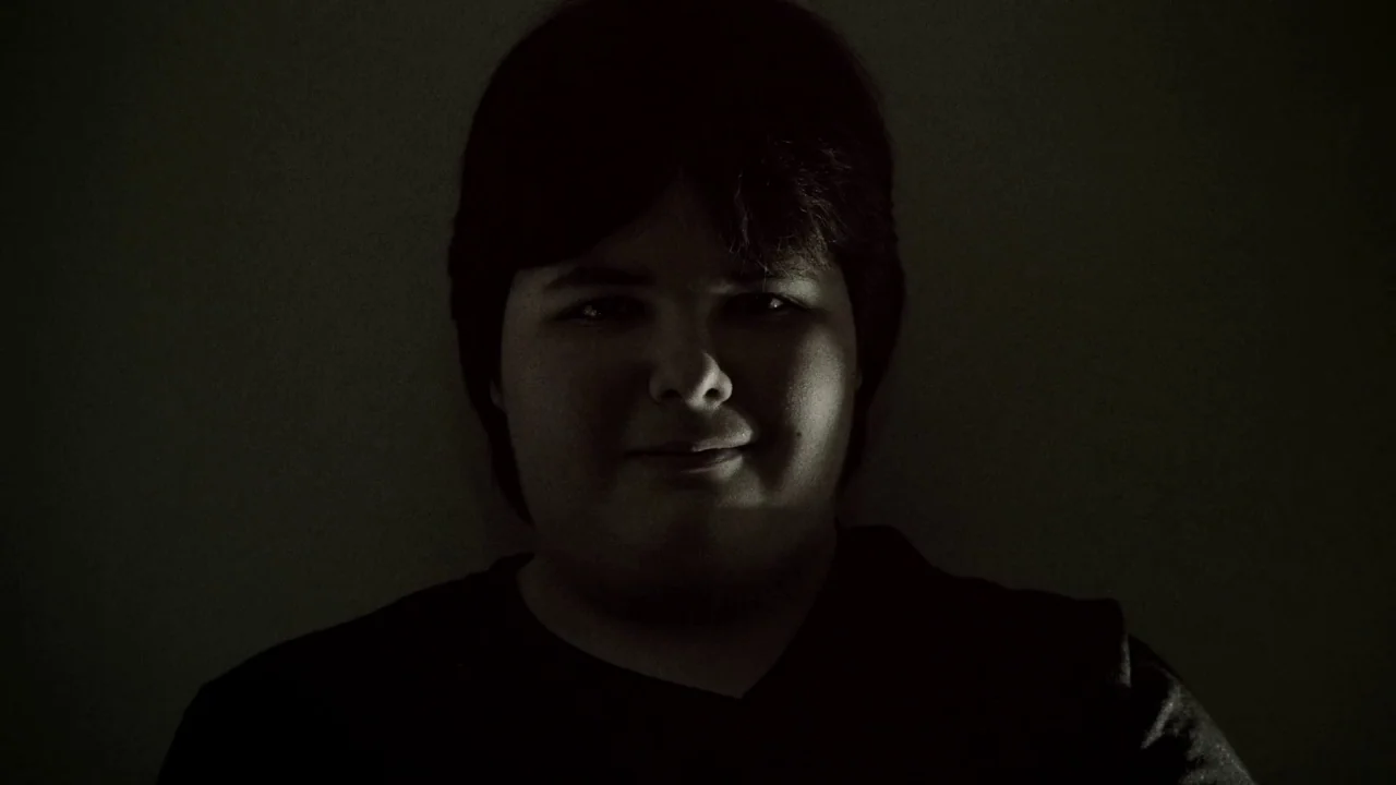 Suburban Gothic (2014) movie still: closeup — A dark indoor setting features a person against a featureless wall. The person stares directly at the…