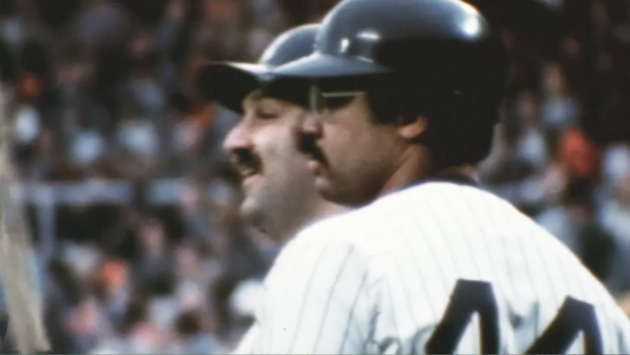 Reggie (2023) movie still: medium — A crowded baseball stadium is visible in the blurred background behind two players. Two men wearing team…