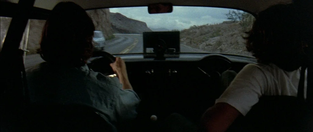 Two Lane Blacktop (1971) movie still: medium — The interior of a moving vintage car shows two people seated in the front. Through the windshield, a…