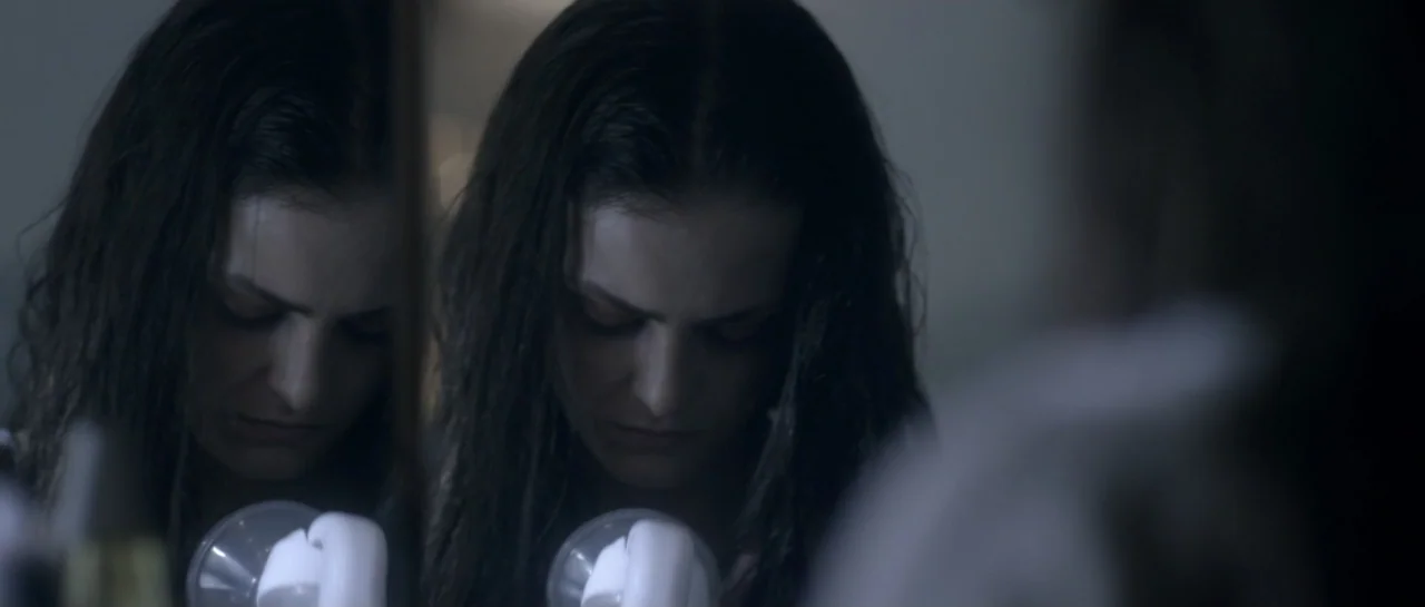 The Lullaby (2019) movie still: closeup — A dimly lit bathroom features a woman positioned in front of a mirror. She looks downward while holding a…