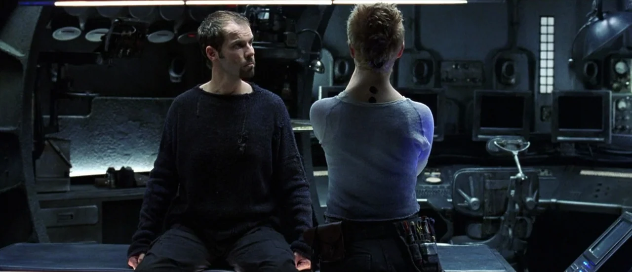 The Matrix Revolutions (2003) movie still: medium — A dimly lit technological station filled with control panels and monitors. A man sits facing a…