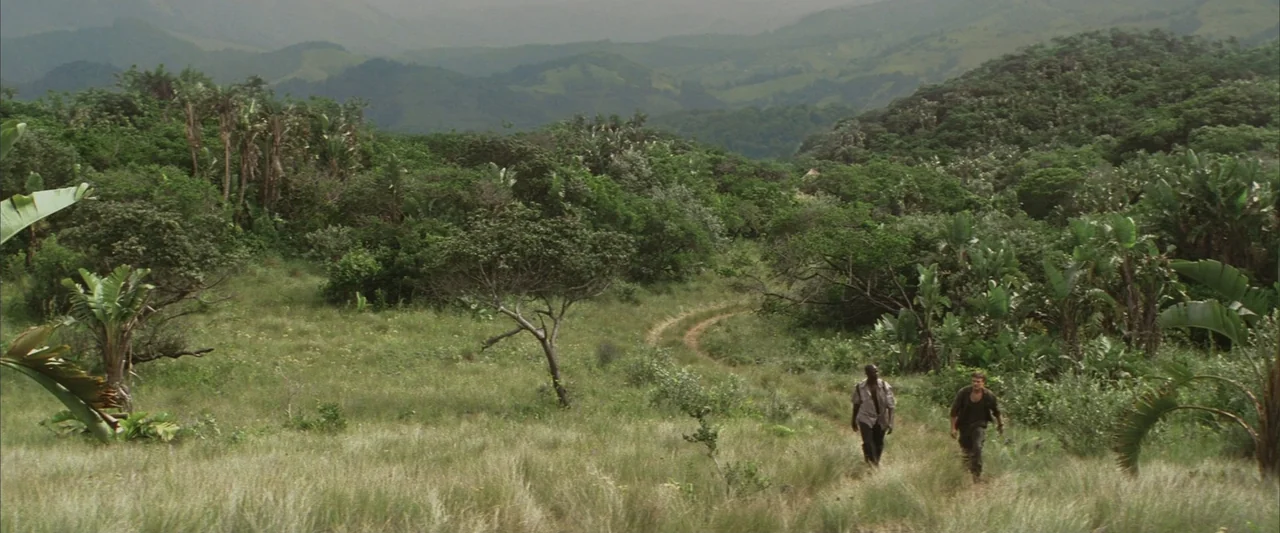 Blood Diamond (2006) movie still: wide — A rolling savanna landscape features dense patches of green vegetation and a worn dirt path. Two men walk…