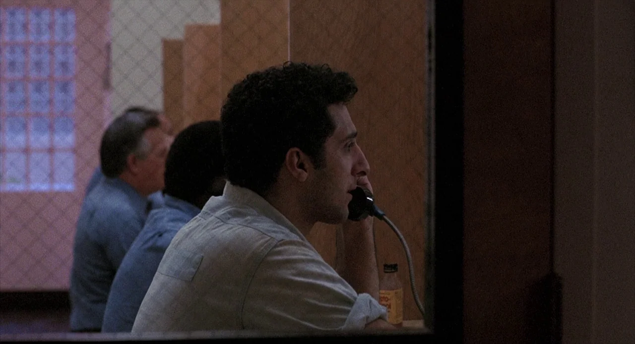 To Live and Die in L.A. (1985) movie still: medium — A man sits behind a wire-mesh divider in a dimly lit visitation area. He holds a telephone…