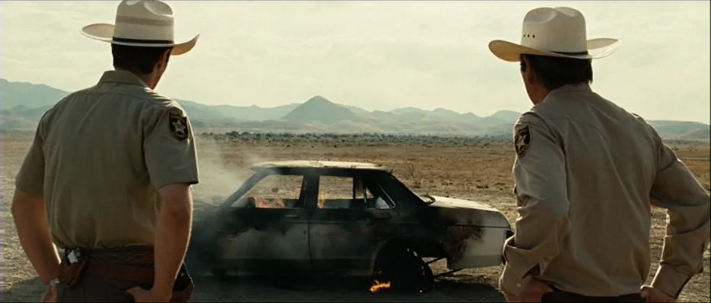 No Country For Old Men movie still: medium — Two law enforcement officers stand in a flat desert expanse under a bright sky. A sedan is engulfed in…