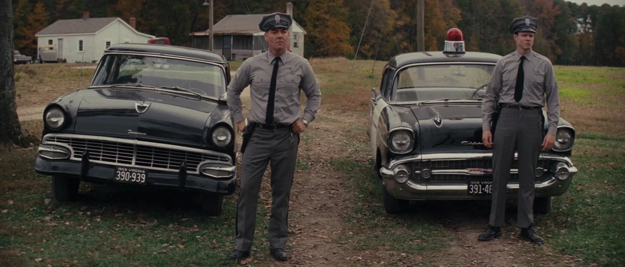 Loving (2016) movie still: fullbody — Two police officers stand in a grassy field in front of vintage patrol cars. The rural background features a…