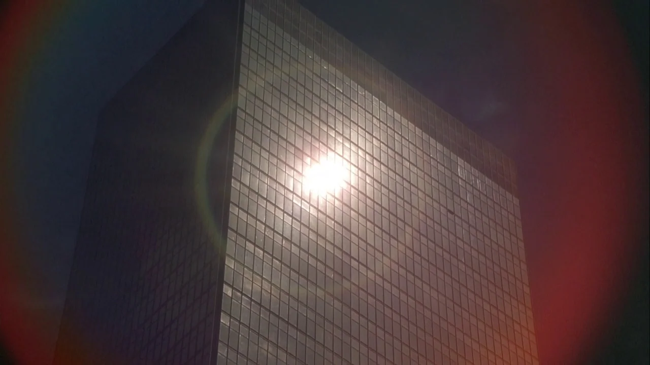 Powaqqatsi (1988) movie still: establishing — A modern glass skyscraper towers into a clear dark sky. The sun beams directly onto the reflective…