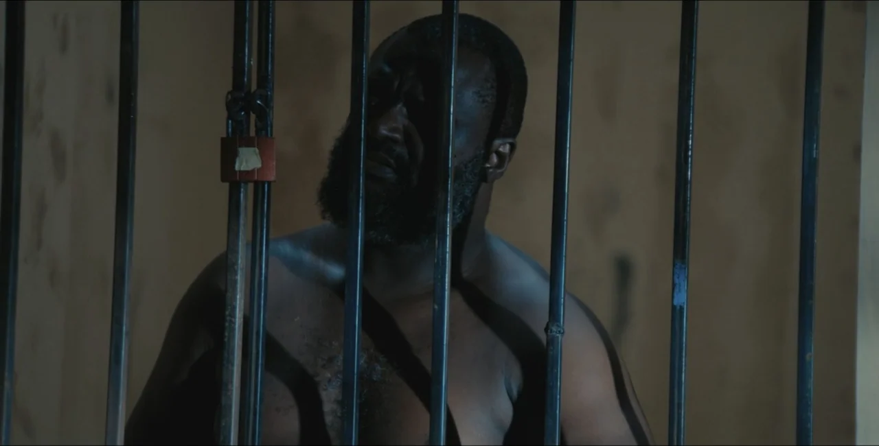 Project Assault (2025) movie still: medium — A dimly lit room features a set of vertical metal bars with a padlock. A shirtless man stands behind…