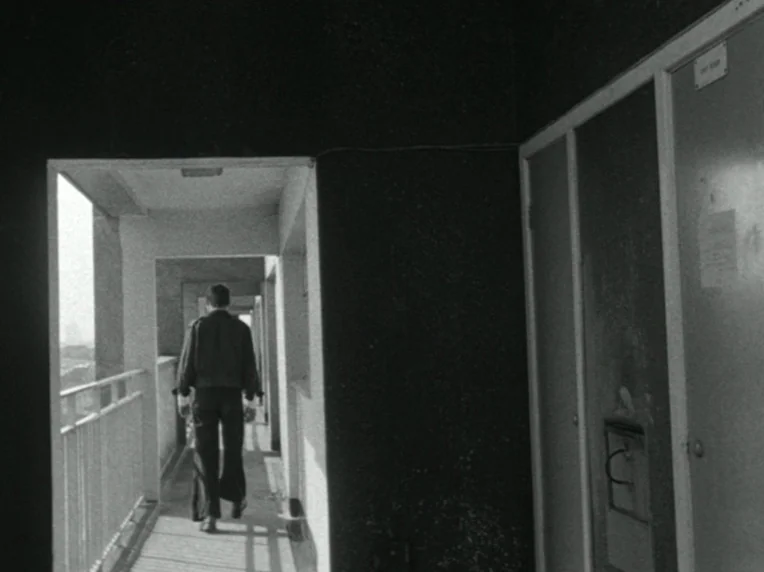 Children (1976) movie still: fullbody — A stark concrete apartment block walkway with heavy shadows and bright sunlight. A man walks away from the…