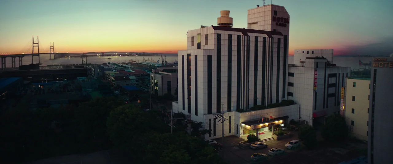 Beasts Clawing At Straws (2020) movie still: establishing — A tall white hotel building stands in a coastal industrial area at sunset. A large…