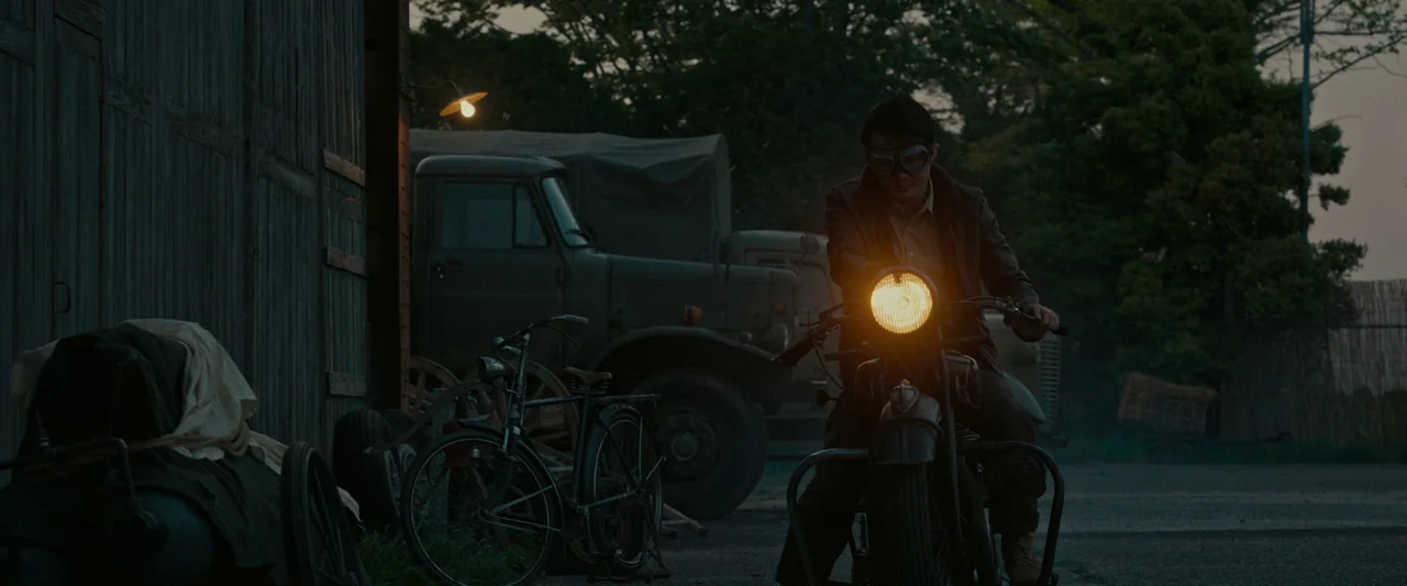 Godzilla Minus One (2023) movie still: medium — A dark outdoor space features a parked truck and bicycles near a wooden structure. A man sits on a…