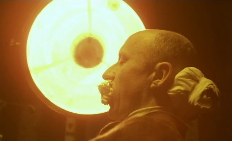 Golem (1980) movie still: closeup — A dark, cramped room features a bright, circular lamp shining directly into the frame. An older man reclines in…