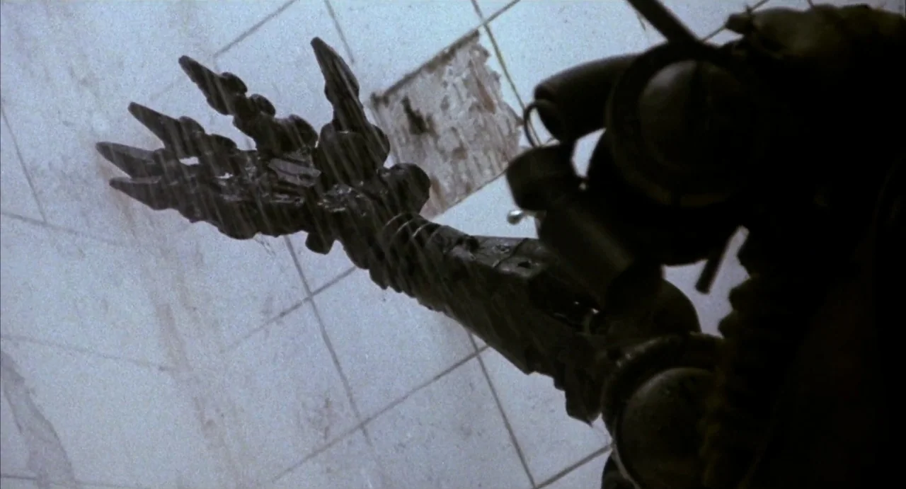 Hardware (1990) movie still: closeup — A dark mechanical appendage reaches out toward a cracked white tiled wall surface. The textured metal claw is…