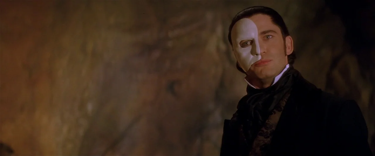 The Phantom of the Opera (2004) movie still: medium — A dark subterranean space features textured rock walls in the background. A man wearing a…