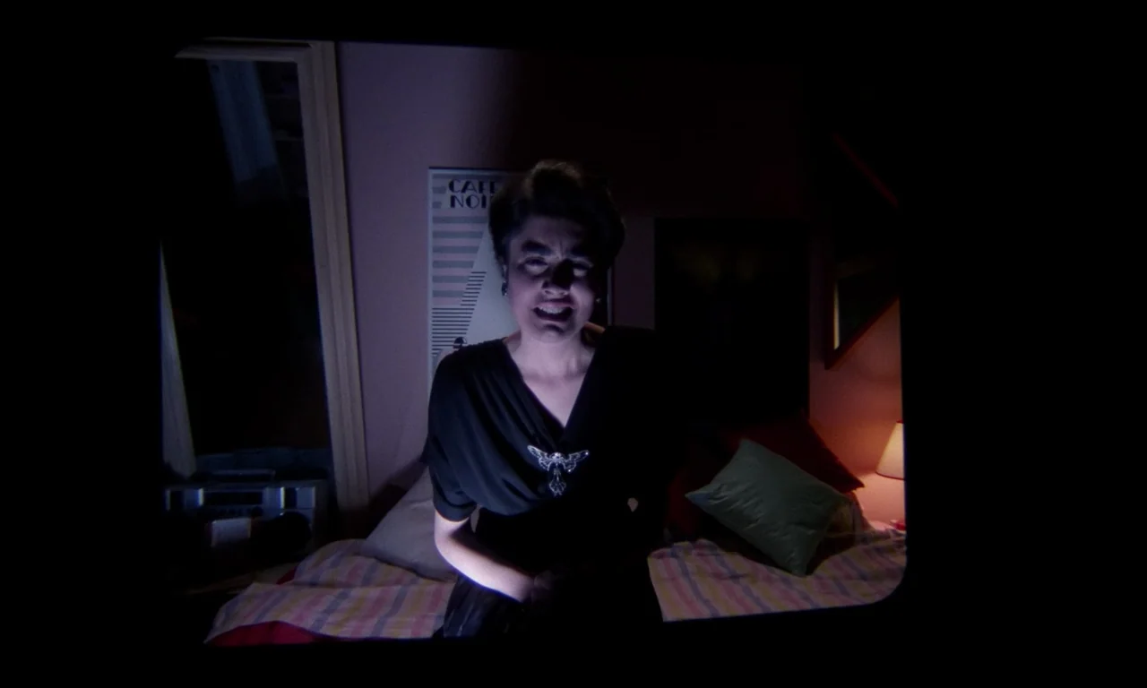 Demons 2 (1986) movie still: medium — A dark bedroom with a bed featuring pillows and a striped blanket. A young woman faces the camera directly…