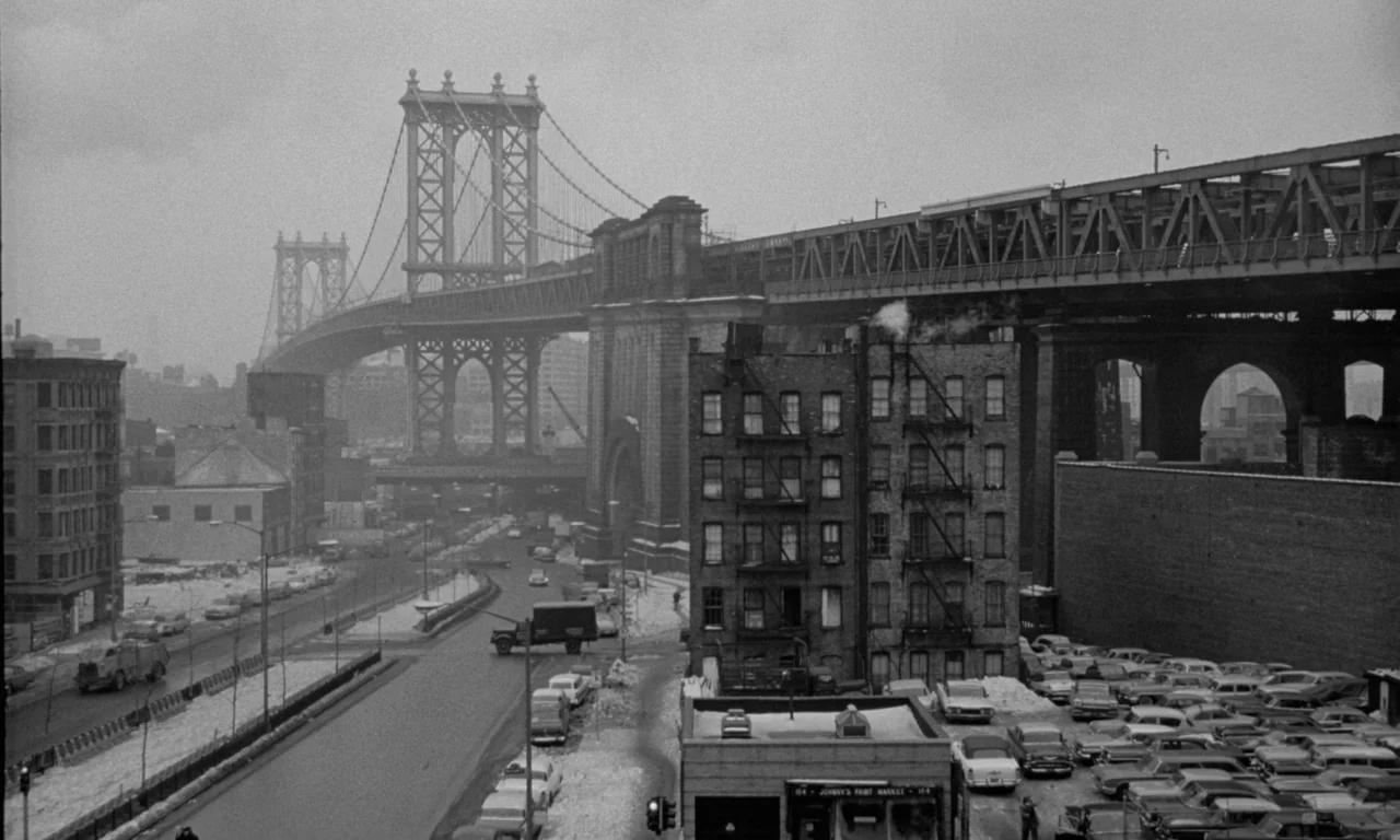 Something Wild (1961) movie still: wide — A dense urban intersection sits beneath a large, elevated iron bridge structure. Rows of parked cars line…