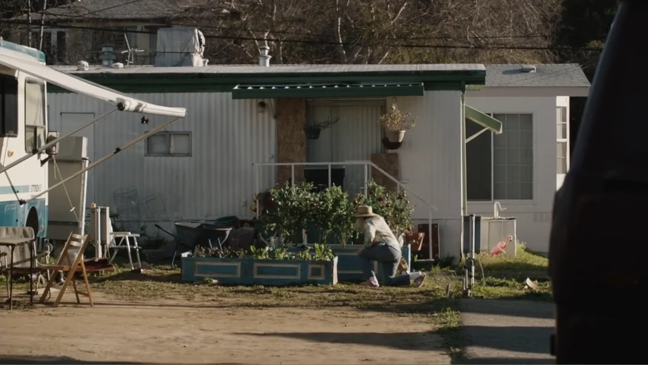 The Cleaner (2021) movie still: wide — A worn mobile home stands behind a makeshift garden bed in a dirt yard. A woman kneels in the grass to prune…