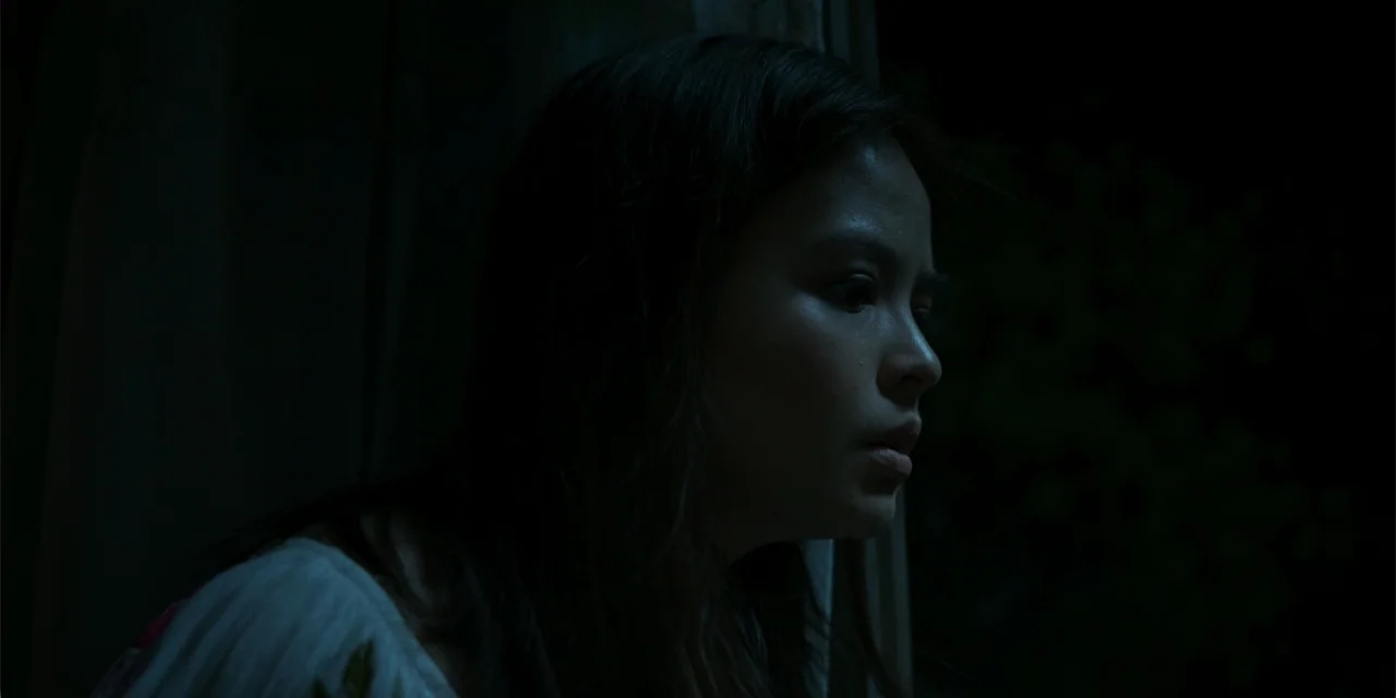 Nokturno (2024) movie still: closeup — A dimly lit room with heavy dark curtains framing the view of a person. A young woman looks intently toward…